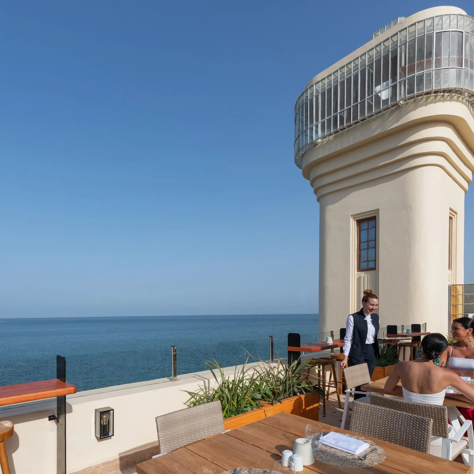 Une terrasse avec des tables et des chaises en plein air, située au bord de la mer, avec un serveur et des clients, devant une tour blanche avec une structure vitrée en haut