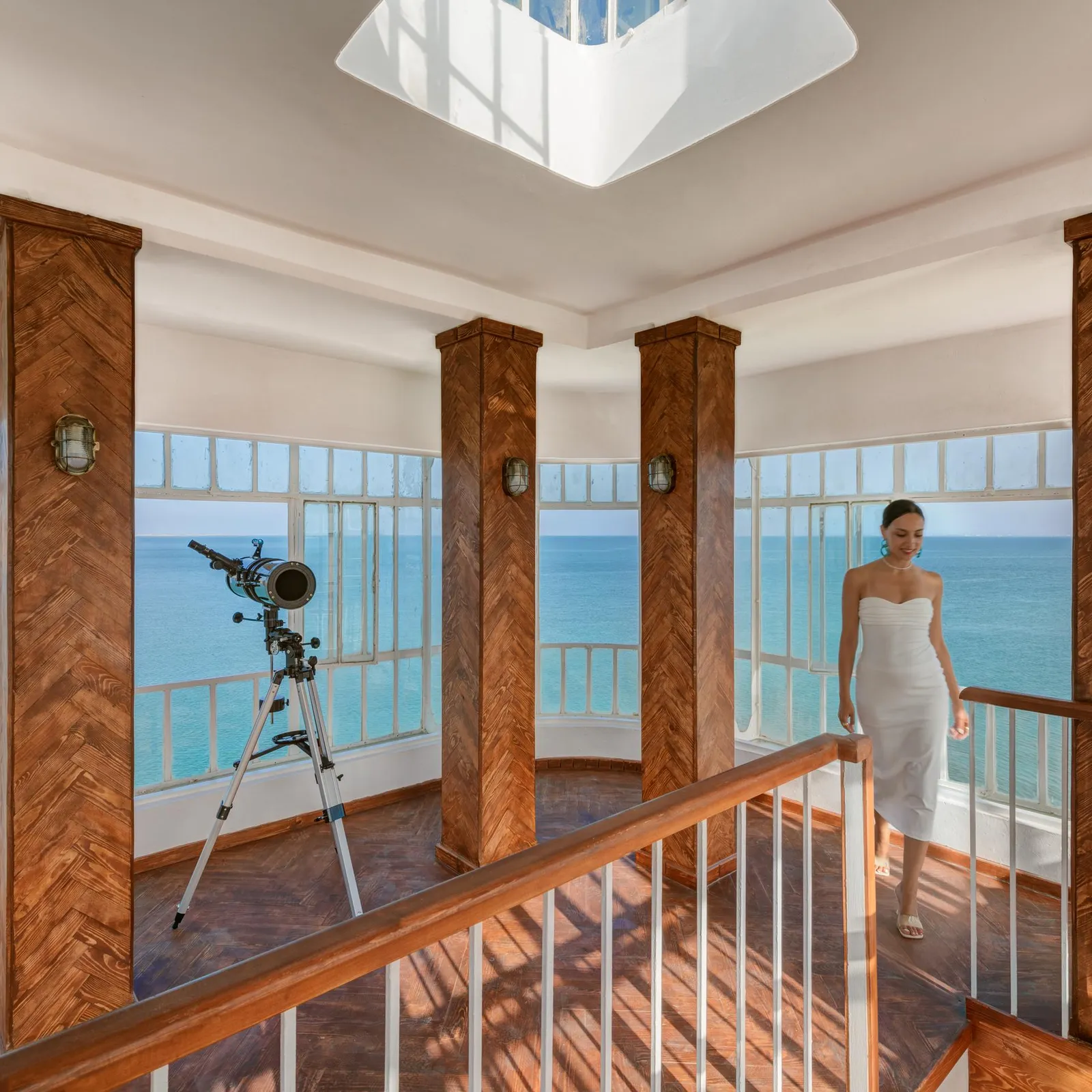 Une femme en robe blanche marche sur un balcon en bois avec vue sur la mer, un télescope est installé près de la rambarde