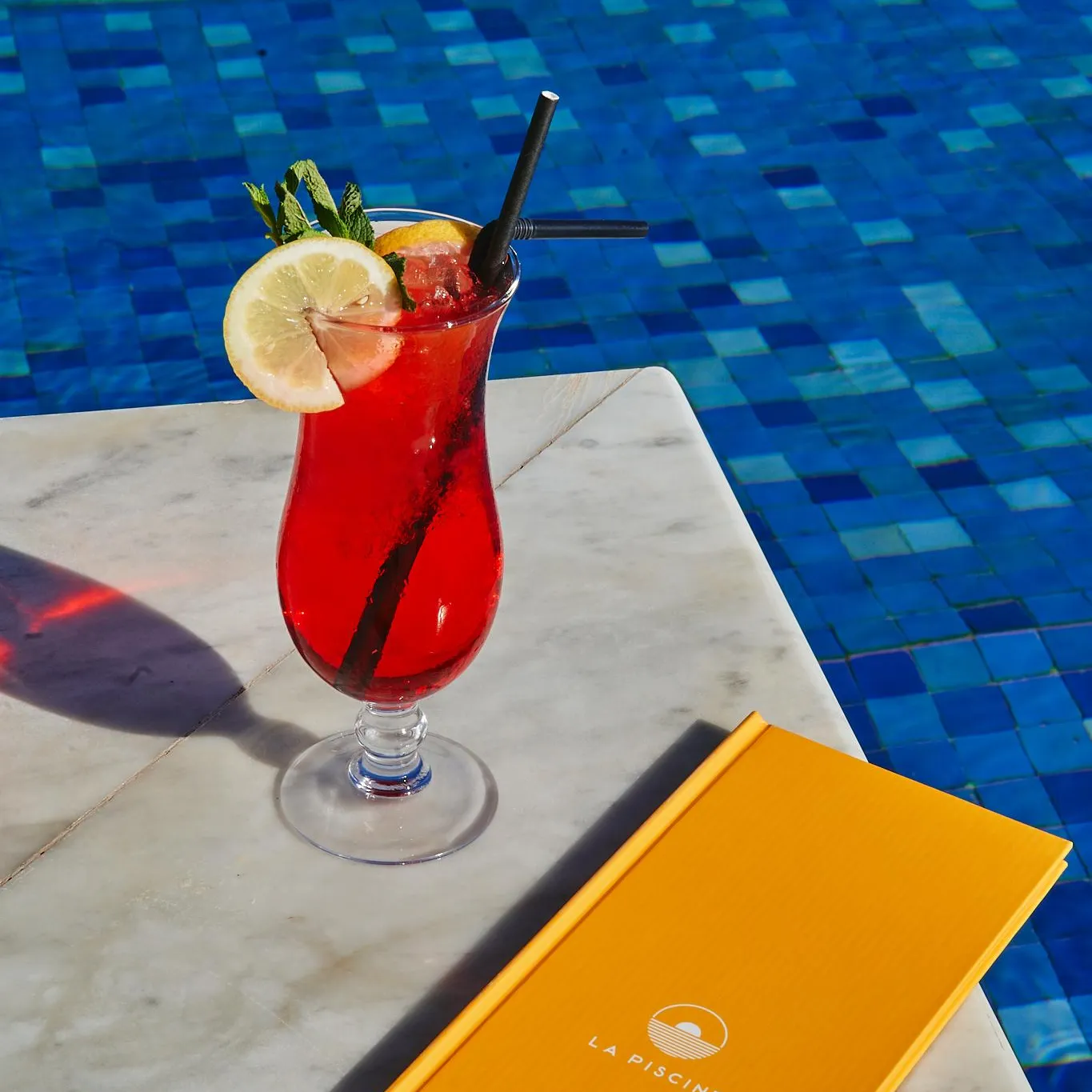 Un verre de boisson rouge avec une tranche de citron et des feuilles de menthe posé sur une table près d'une piscine, à côté d'un menu jaune