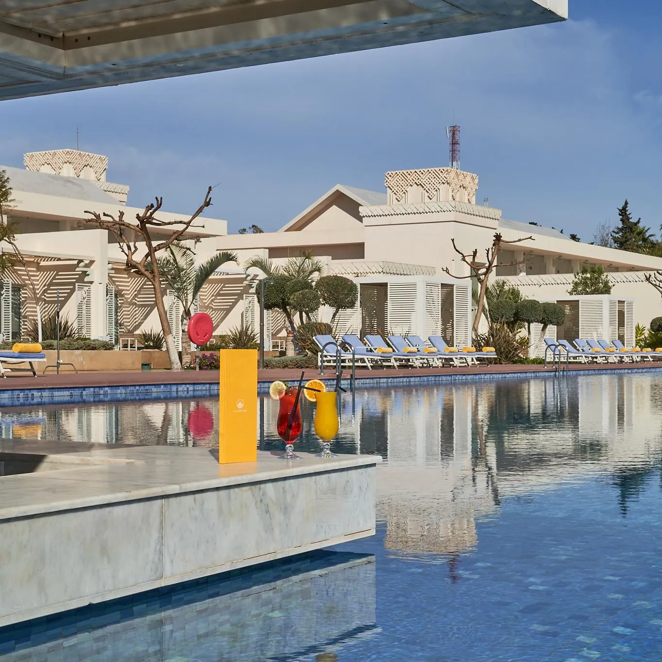 Piscine avec deux verres de boissons colorées sur le bord, entourée de transats et de bâtiments blancs avec des toits en terrasse