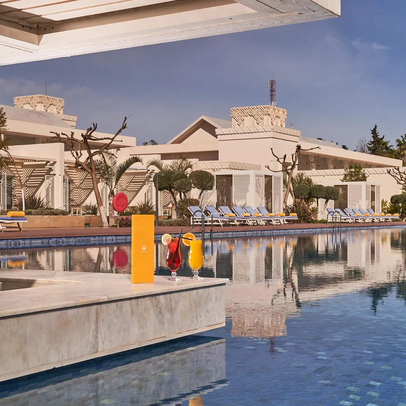 Deux verres de boissons colorées sur une table près d'une piscine avec un bâtiment beige en arrière-plan