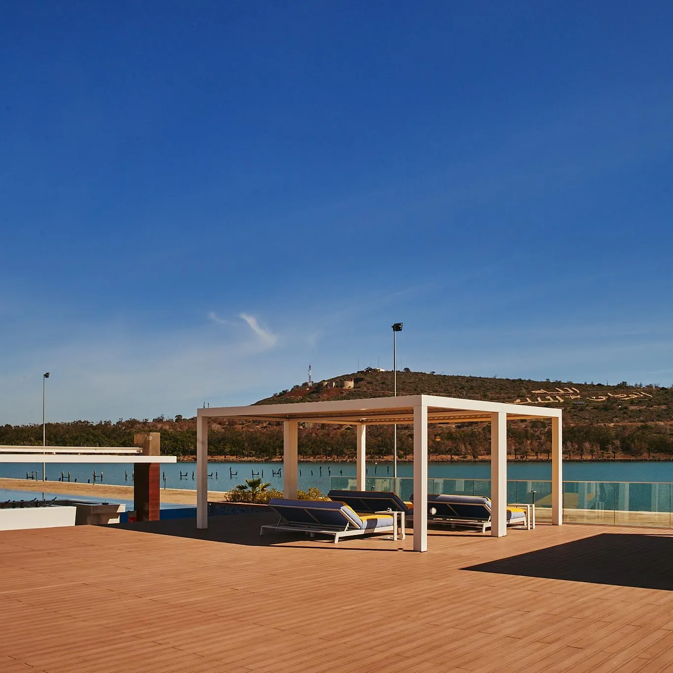 Pérgola blanche avec transats sur une terrasse en bois près d'une piscine et d'un plan d'eau avec des collines en arrière-plan