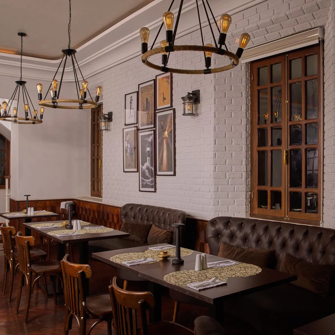 Restaurant avec tables en bois, chaises et banquettes en cuir, éclairé par des suspensions à ampoules nues, murs en briques blanches et cadre en bois aux fenêtres