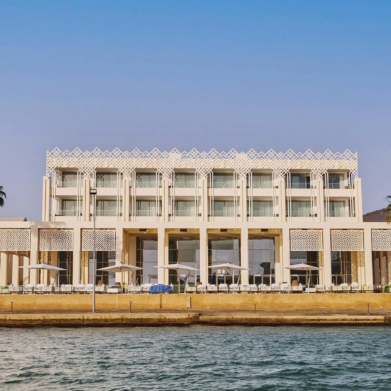 Un bâtiment blanc de style moderne avec des balcons et des colonnes, situé au bord de l'eau avec des palmiers et des tables sous des parasols