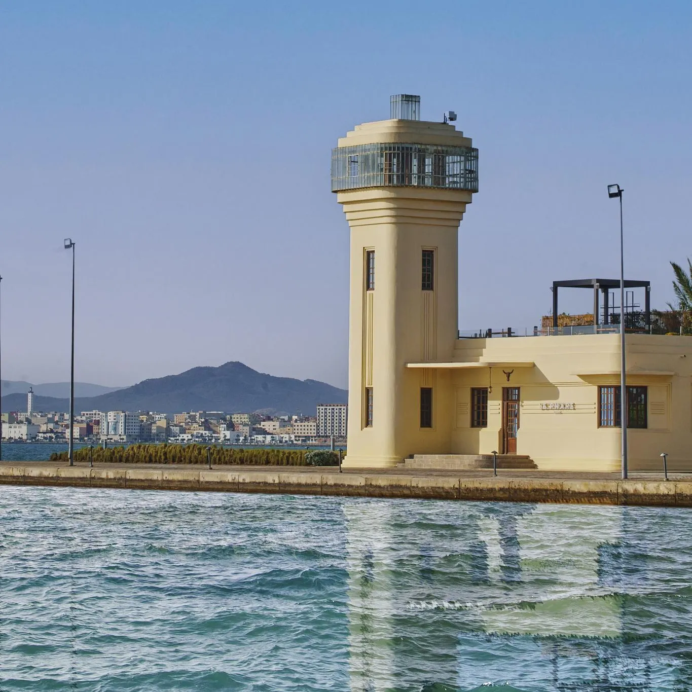 Tour de contrôle jaune avec un observatoire en haut, située au bord de l'eau avec des bâtiments et des montagnes en arrière-plan
