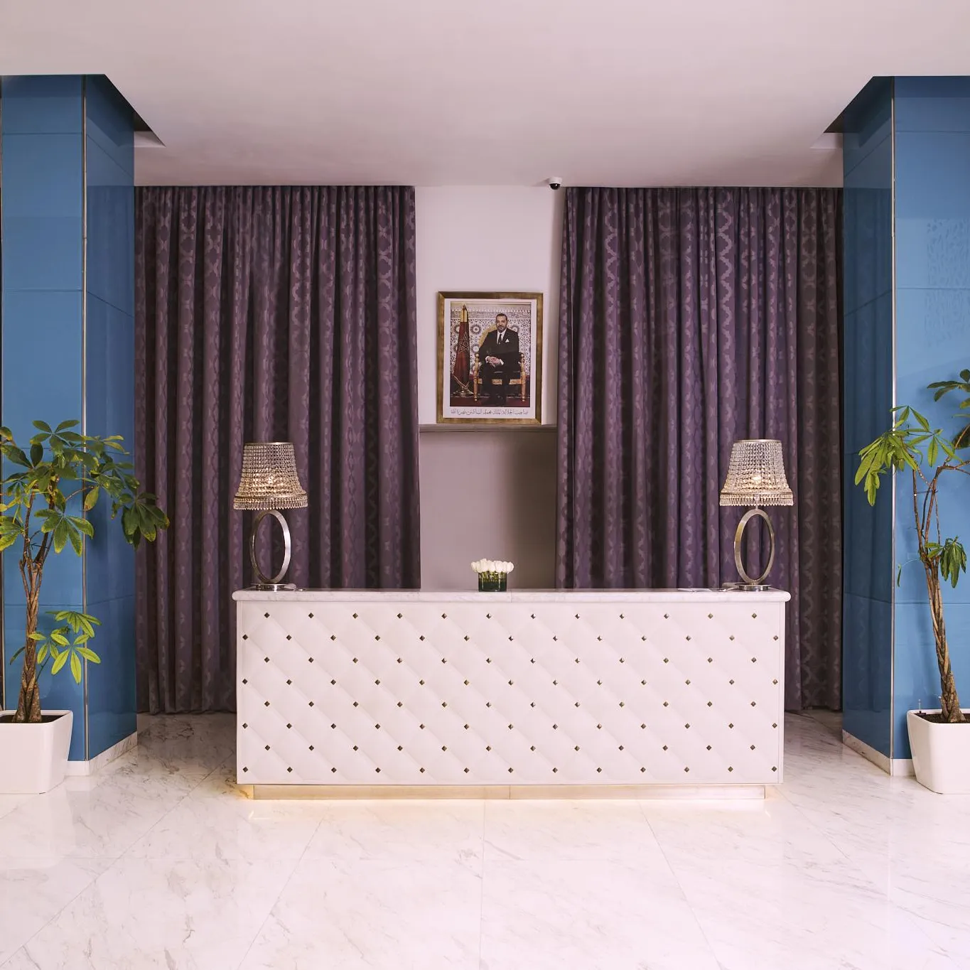 Réception d'hôtel avec un comptoir blanc, deux lampes et trois plantes vertes devant des rideaux violets