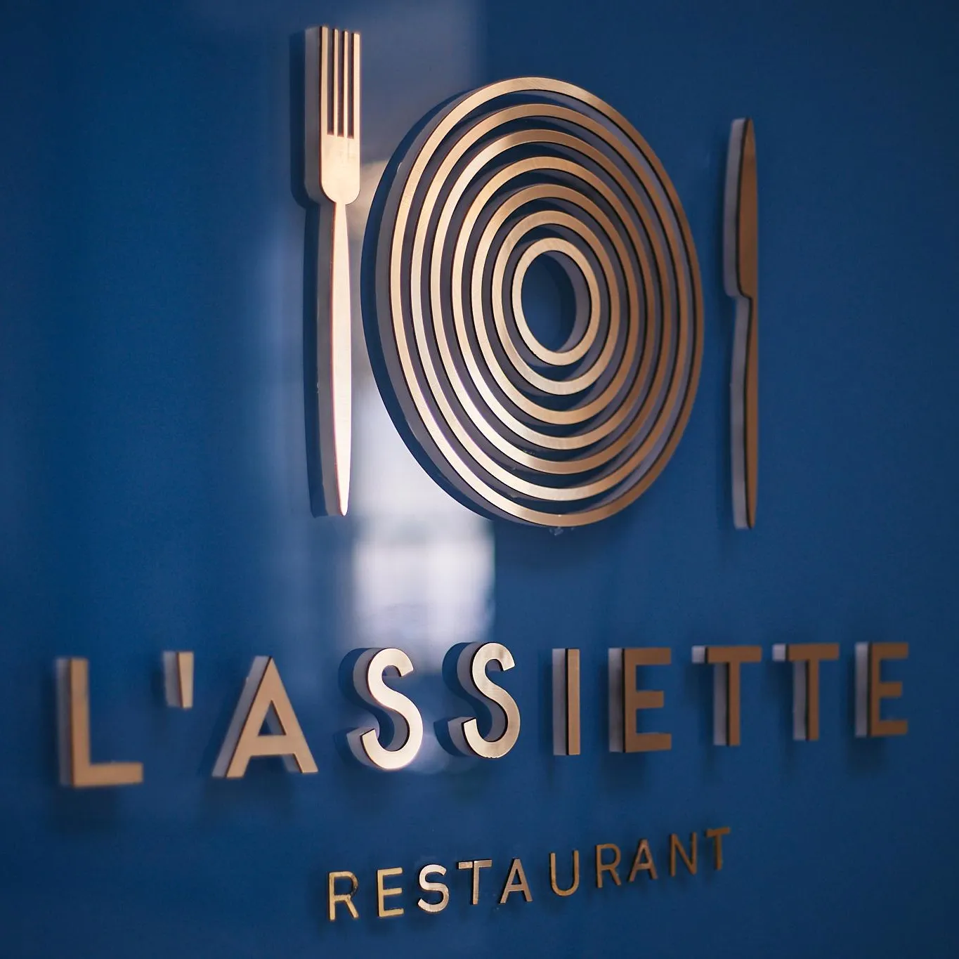 Plaque murale d'un restaurant nommé "L'Assiette" avec un logo représentant une assiette entre deux couverts