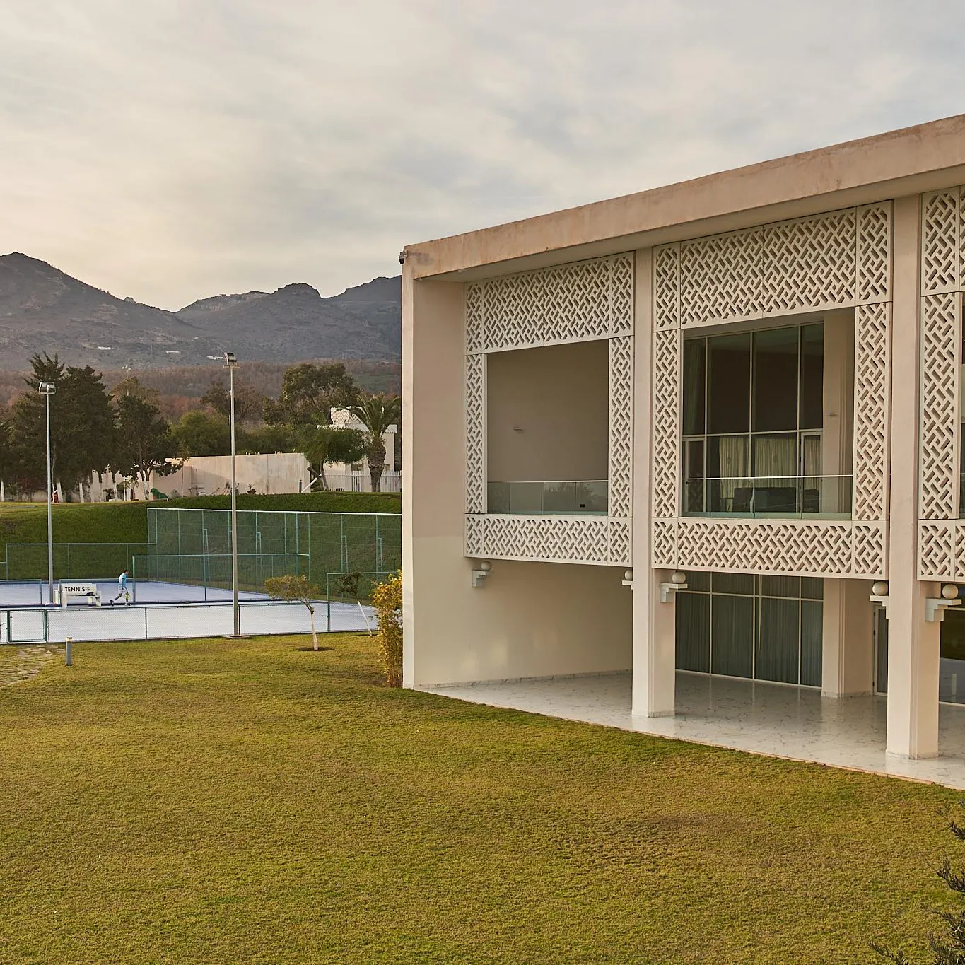 Bâtiment moderne avec façade à motifs géométriques, entouré d'une pelouse et de courts de tennis, avec des montagnes en arrière-plan