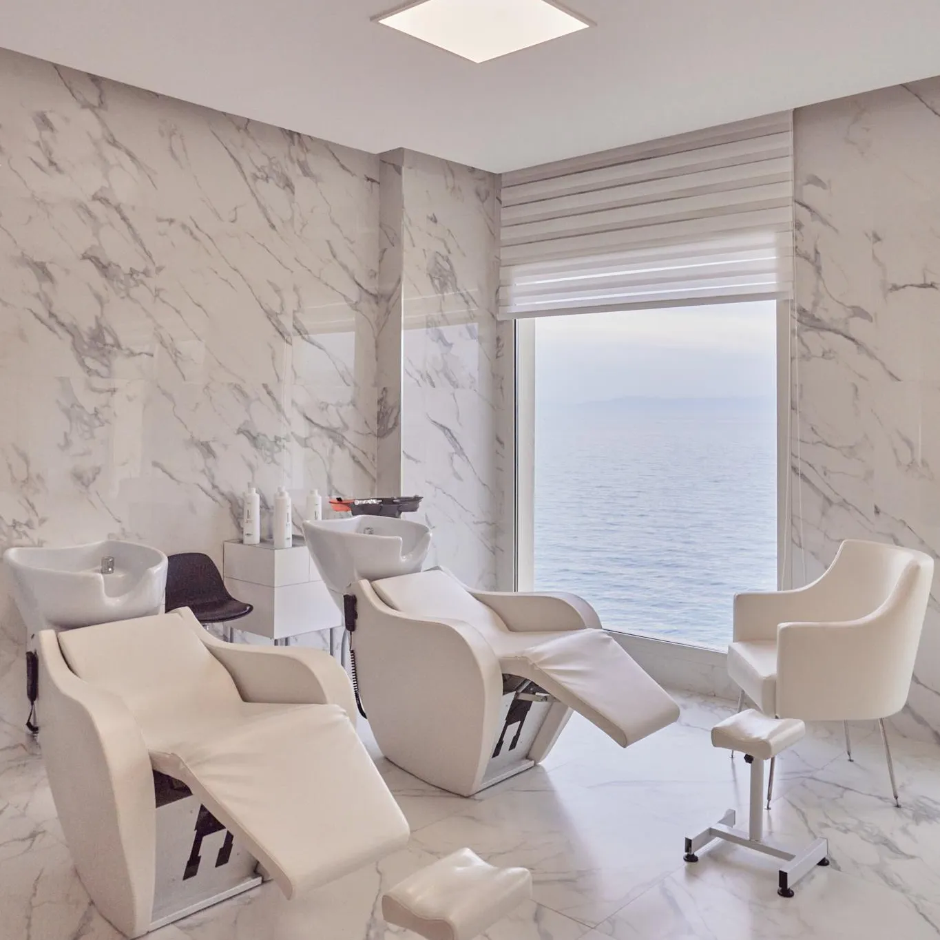 Salon de coiffure avec des fauteuils de coiffure blancs et des murs recouverts de marbre blanc, donnant sur une vue sur la mer à travers une grande fenêtre
