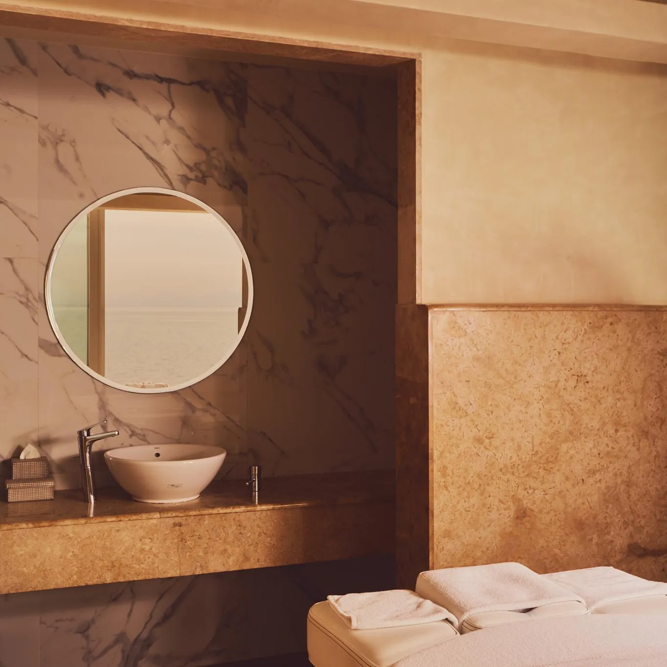 Un lavabo encastré dans un plan de travail en pierre avec un miroir rond au-dessus, à côté d'une table de massage recouverte de serviettes blanches