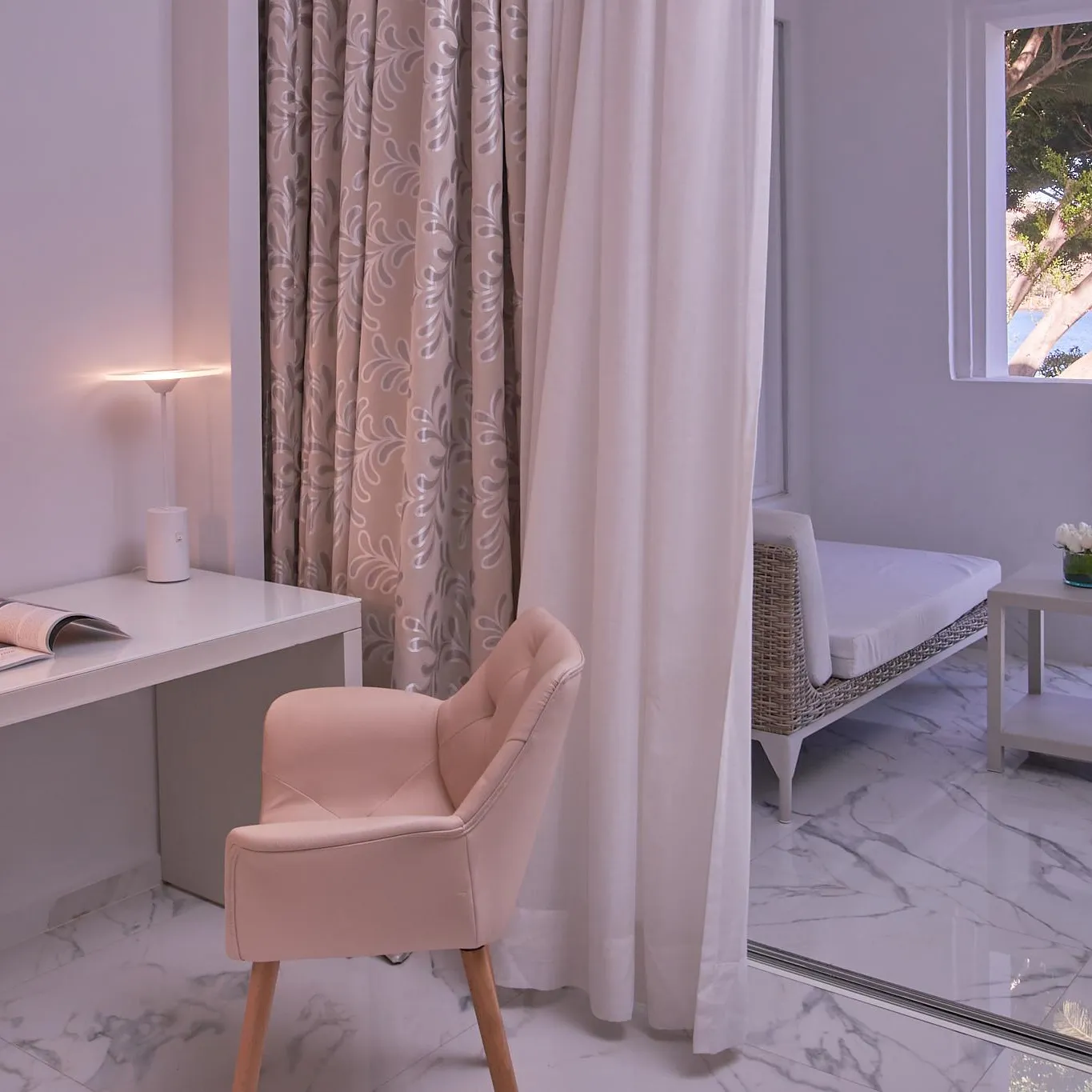 Bureau blanc avec chaise rose, lampe allumée, magazines et décor floral près d'une fenêtre donnant sur un jardin avec des arbres