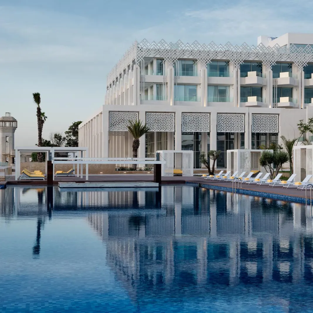 Piscine à débordement devant un hôtel moderne avec des transats et des palmiers, vue sur la mer en arrière-plan
