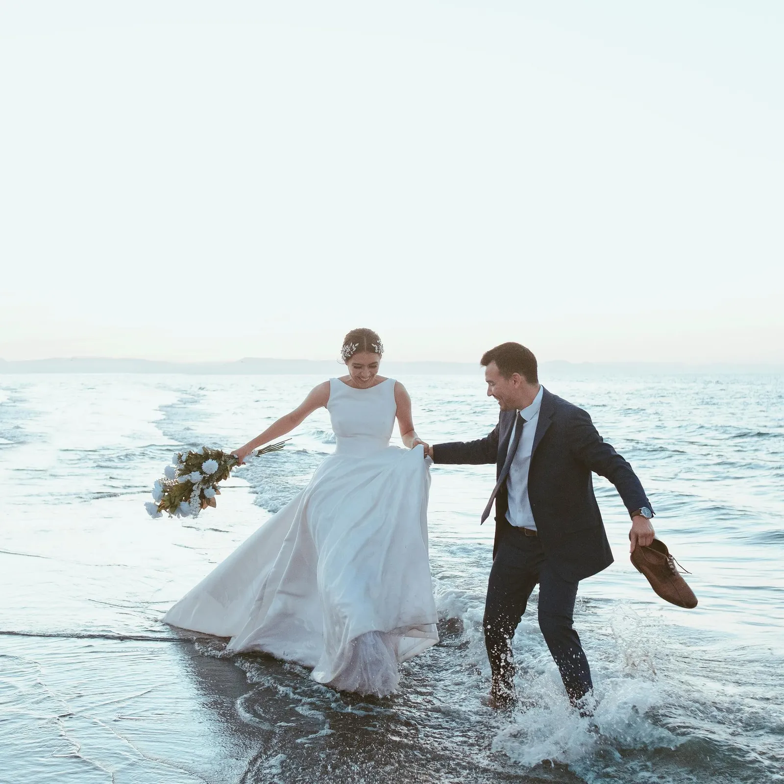 Un couple marche pieds nus dans l'eau, la mariée tenant un bouquet et l'homme tenant ses chaussures