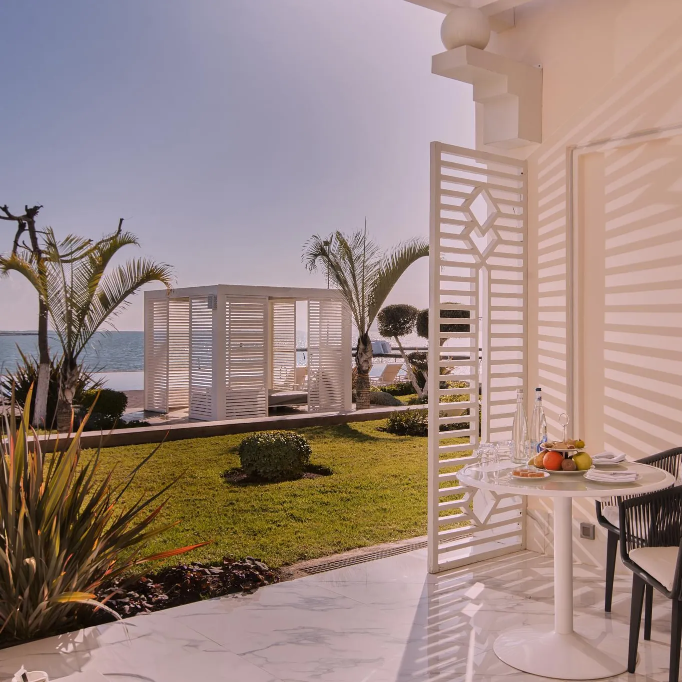 Terrasse avec table, chaises et plantes, vue sur la mer avec un bâtiment blanc en arrière-plan