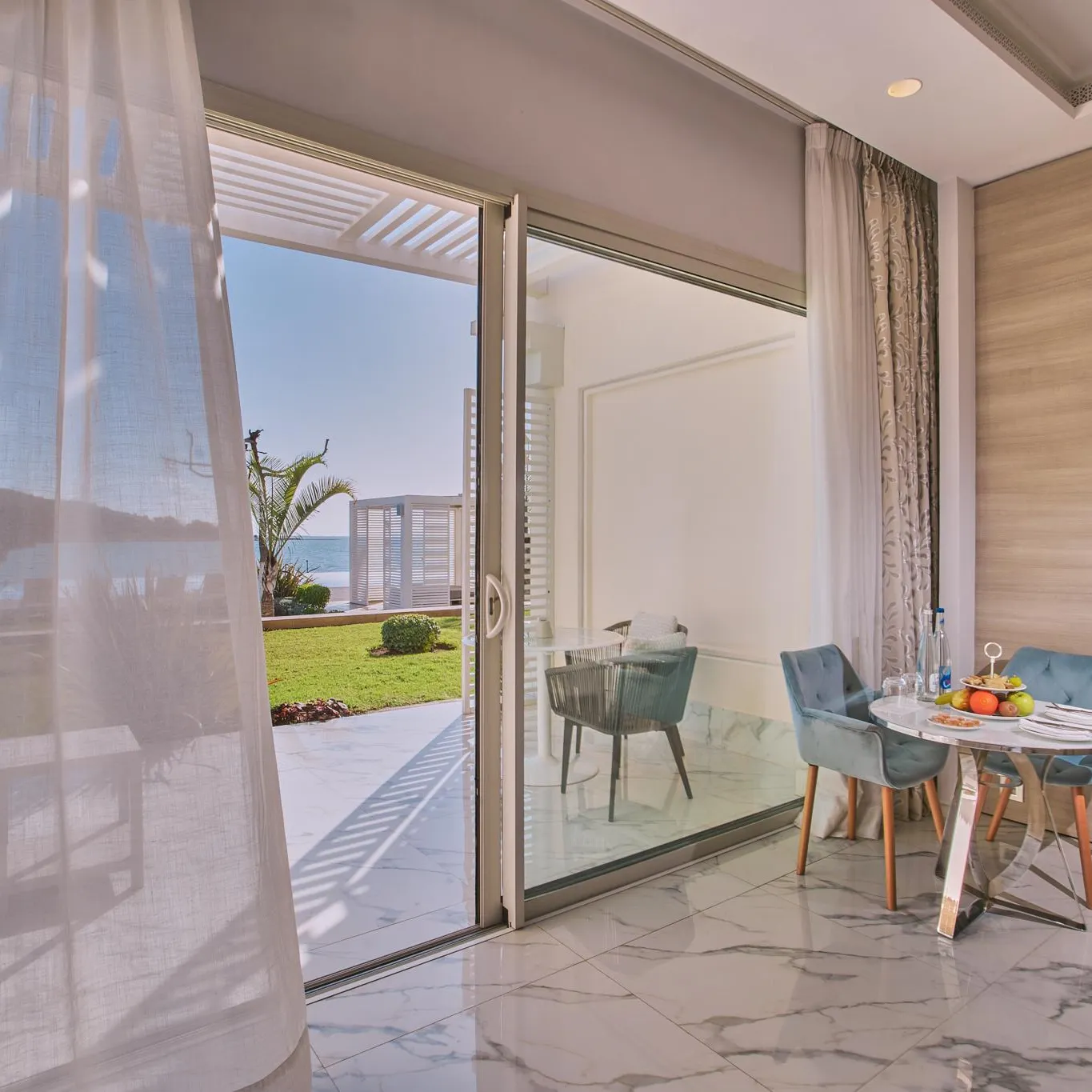 Une table avec des fruits et des verres est installée près d'une porte vitrée ouverte sur un balcon avec vue sur la mer
