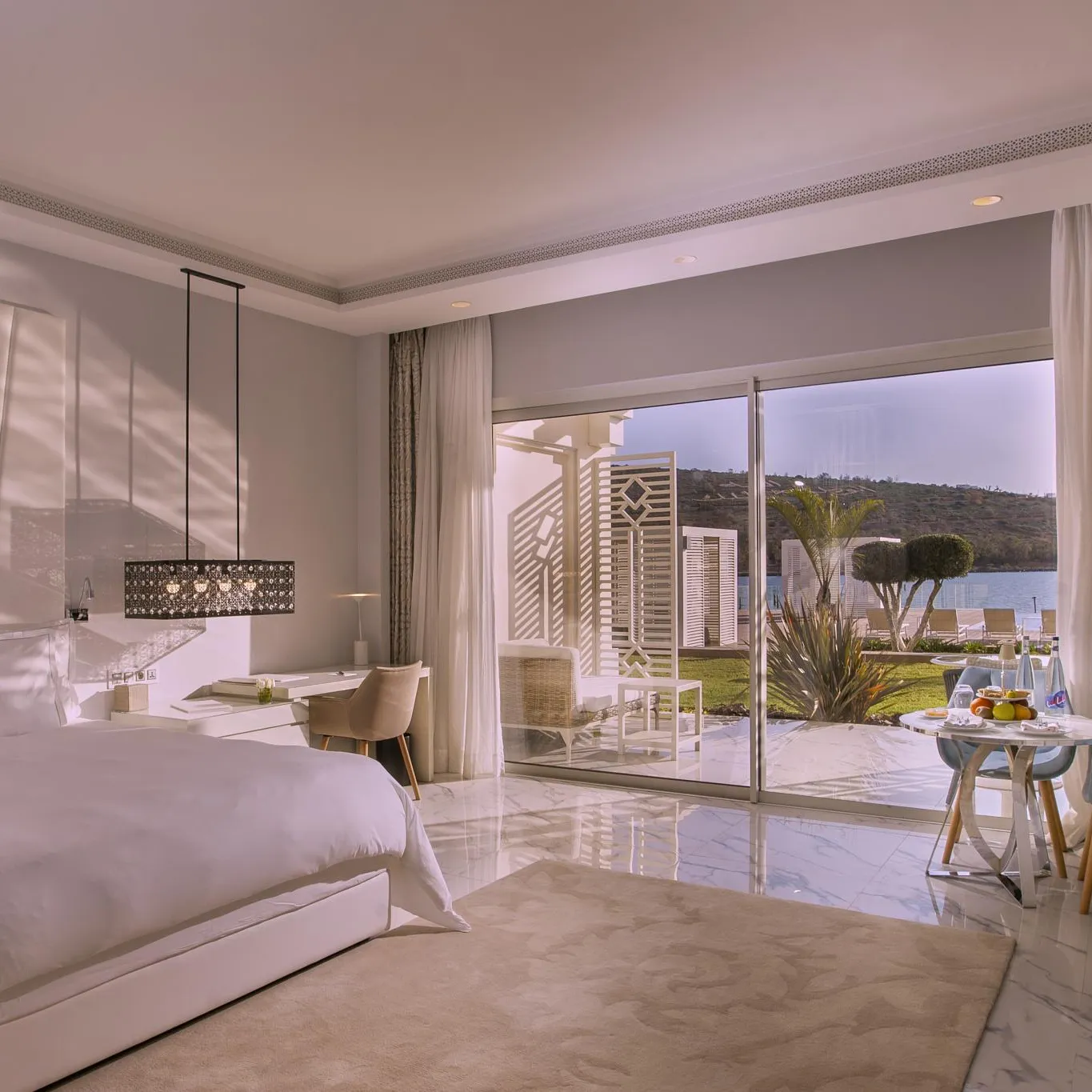 Chambre luxueuse avec lit blanc, table de travail, et vue sur une terrasse et un paysage extérieur depuis de grandes baies vitrées