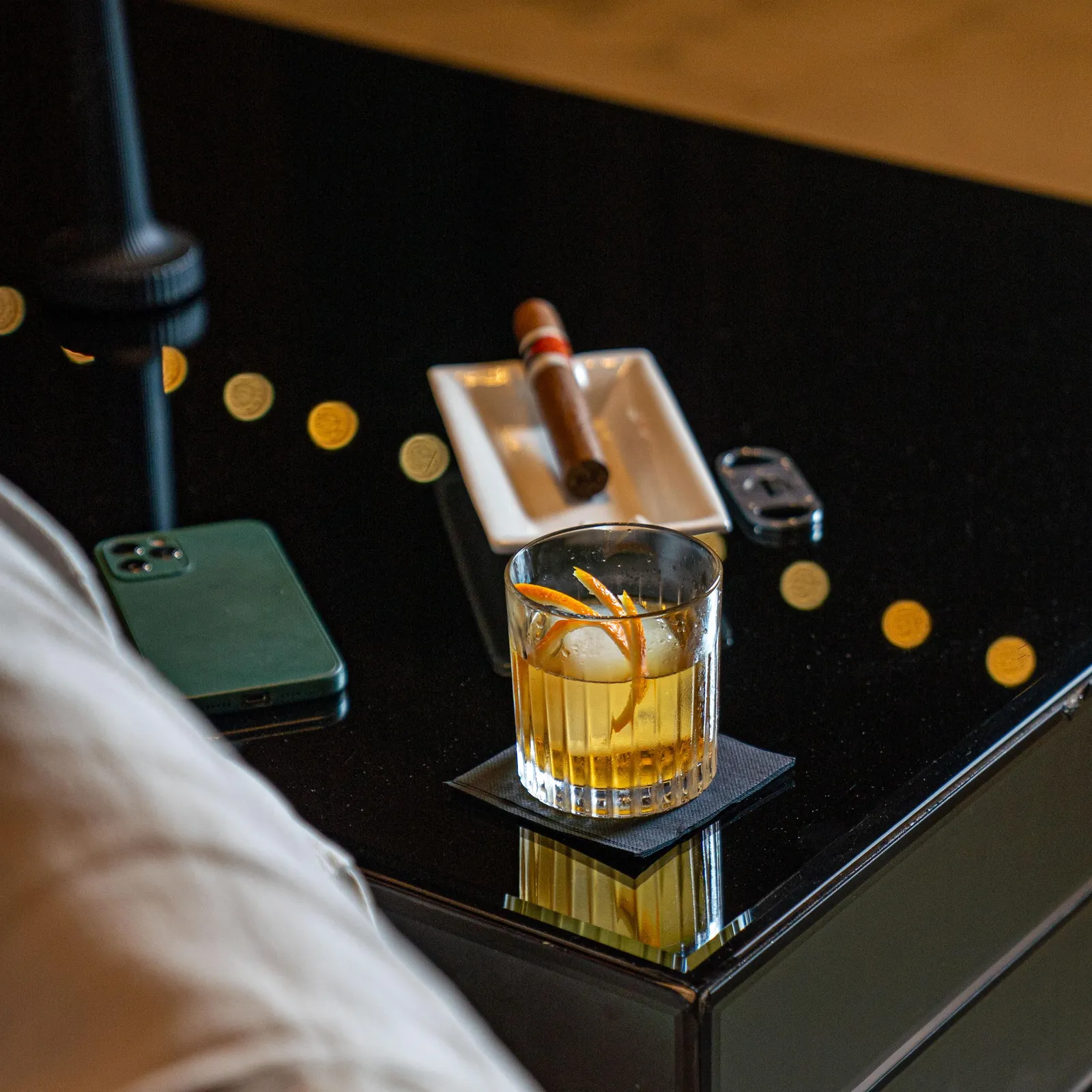 Un verre de cocktail avec des glaçons et une tranche d'orange sur une table, à côté d'un téléphone portable et d'un étui à cigares