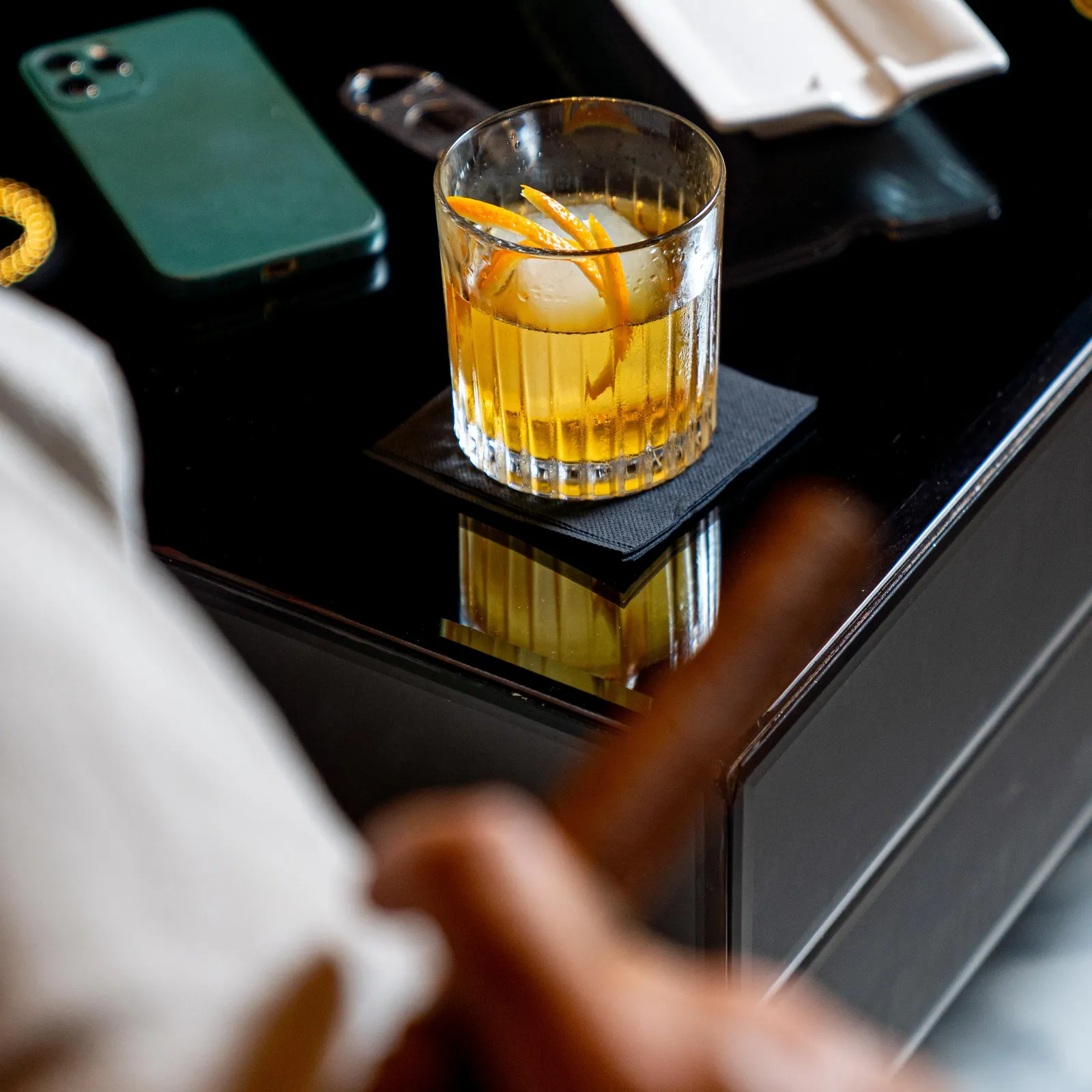 Un verre contenant un liquide jaune avec des glaçons et une tranche d'orange est posé sur une table noire, à côté d'un téléphone portable vert