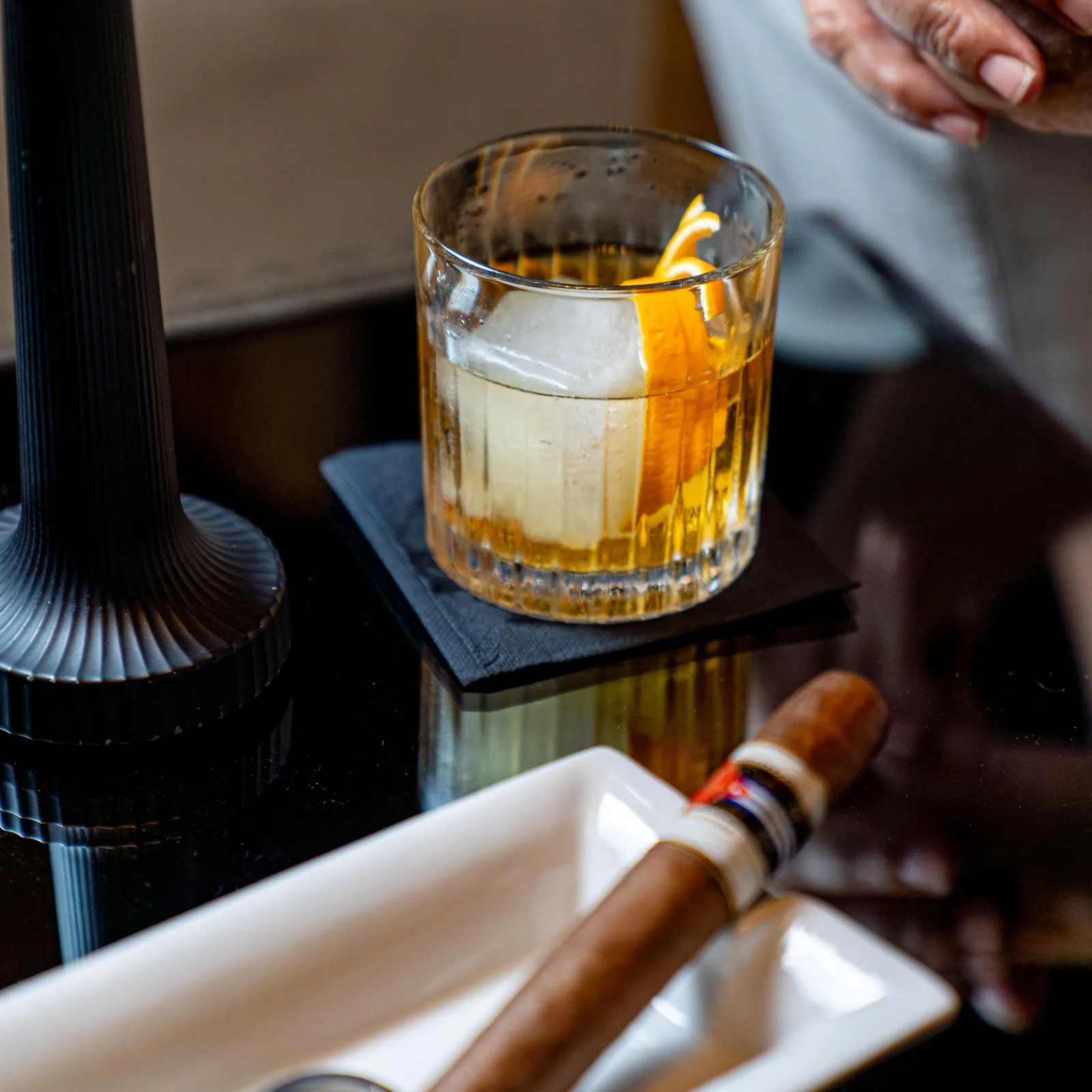 Un verre contenant un liquide ambré avec des glaçons et une tranche d'agrumes, un cigare dans un étui et des mains posées sur une table