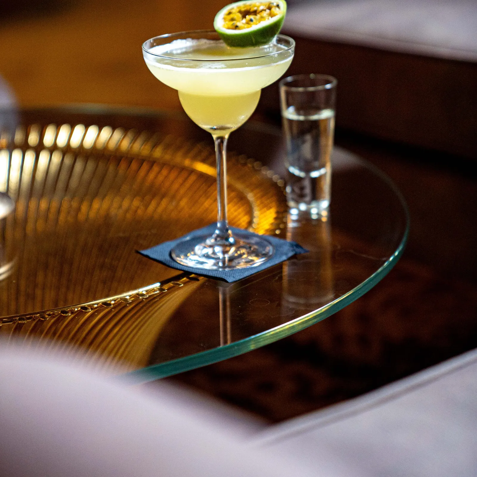 Un verre à cocktail contenant une boisson claire avec une tranche de fruit posé sur le bord, accompagné d'un petit verre d'eau, sur une table en verre