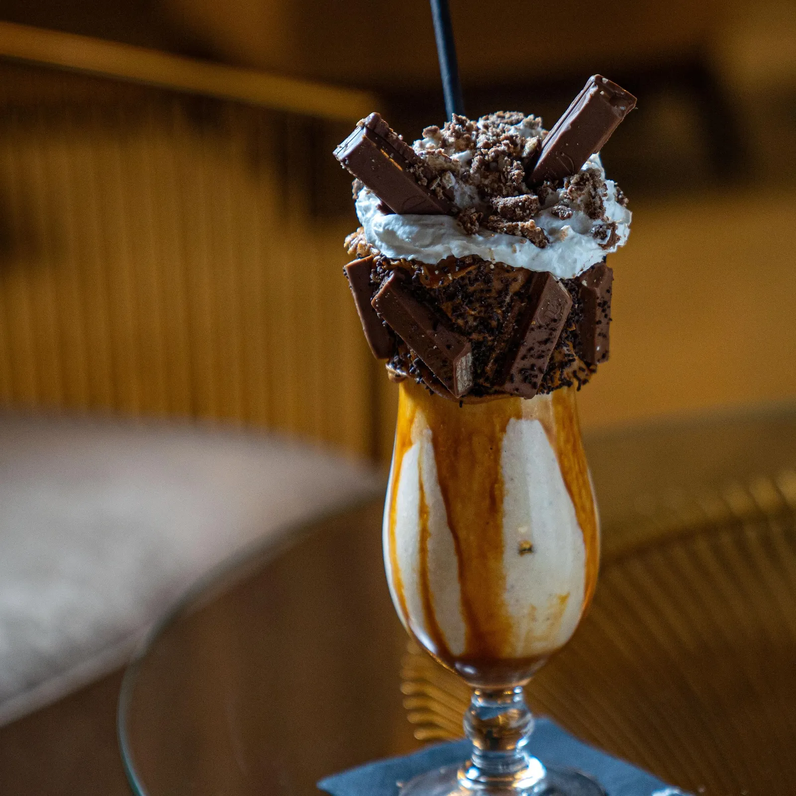 Un verre transparent contient une boisson avec une garniture de crème chantilly, de morceaux de biscuit et de chocolat, posé sur une soucoupe métallique