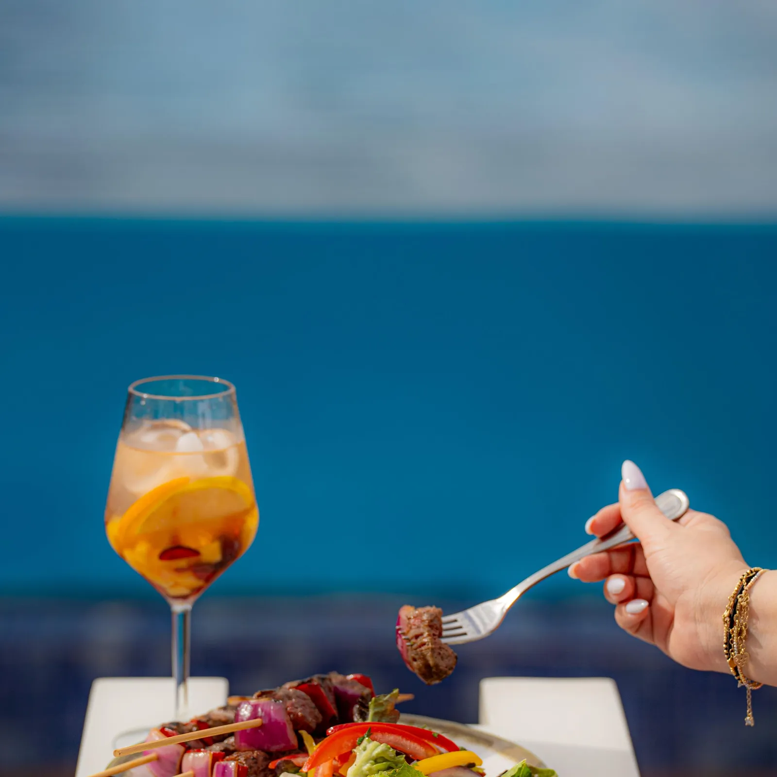 Une main tient une fourchette avec un morceau de viande au-dessus d'une assiette de brochettes et de salade, avec un verre de boisson à l'orange en arrière-plan devant un ciel bleu
