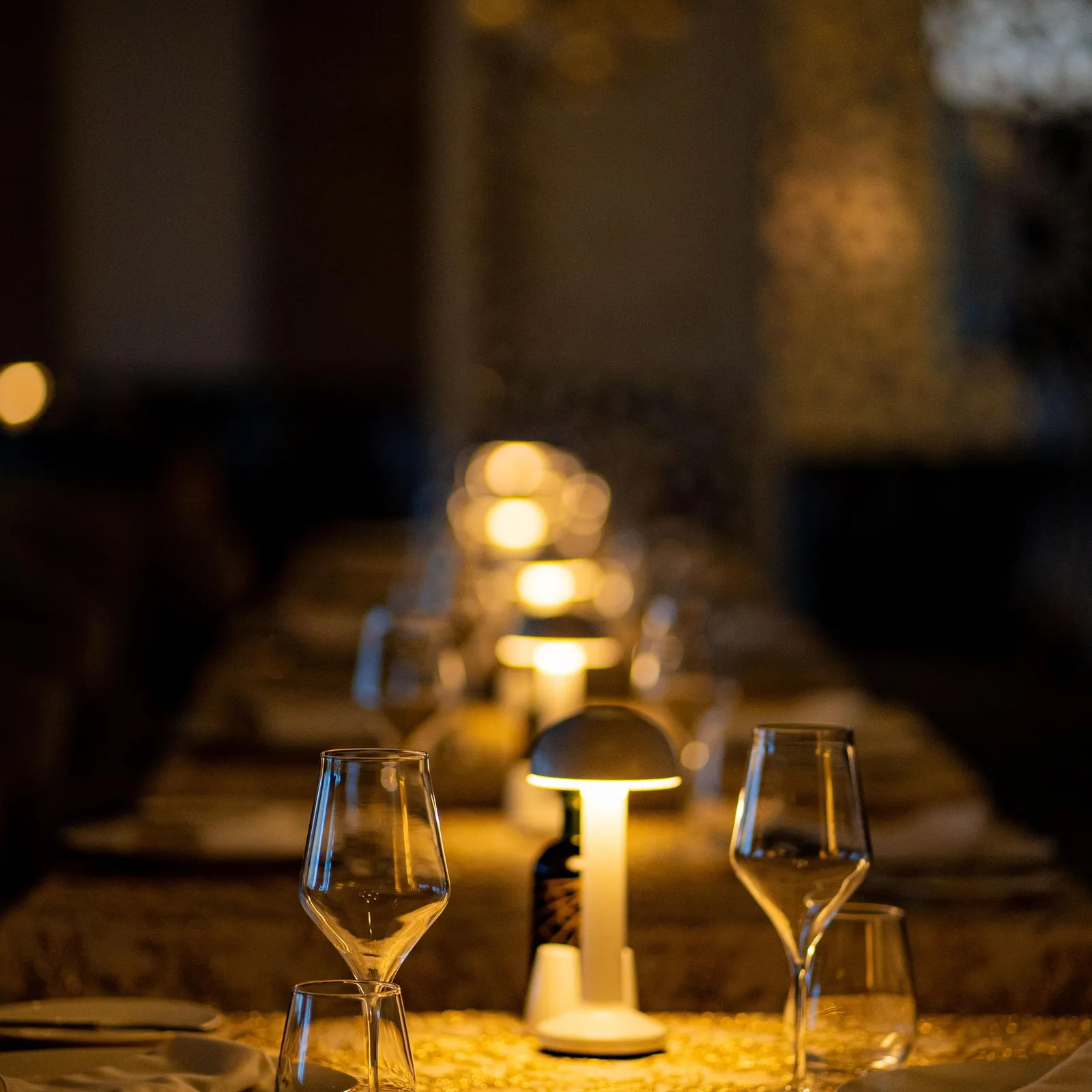 Table dressée avec verres à vin, nappe et bougies, éclairée par des lampes en fond