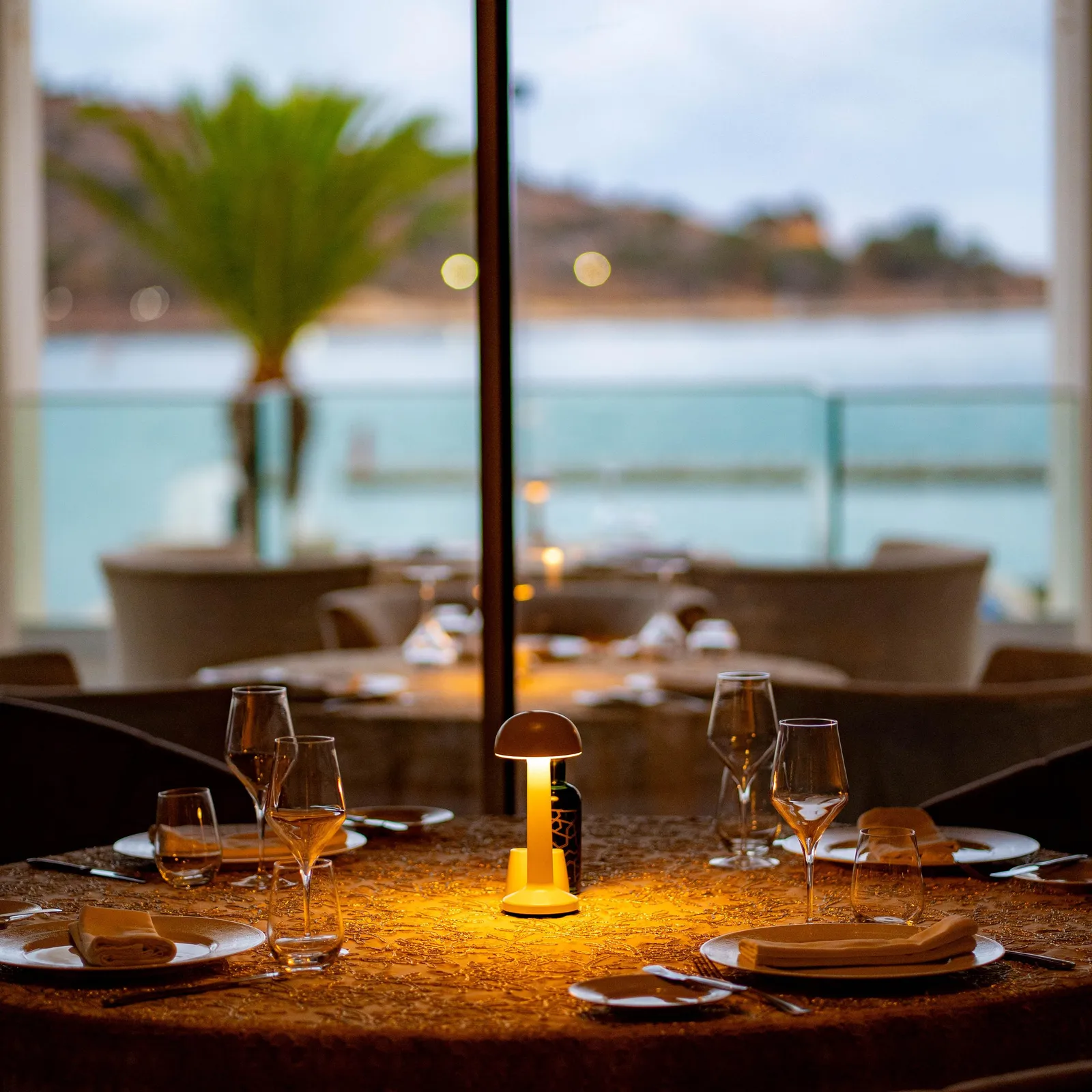 Table dressée avec verres à vin et bougie allumée, vue à travers une fenêtre donnant sur une terrasse et un paysage extérieur avec palmiers et mer