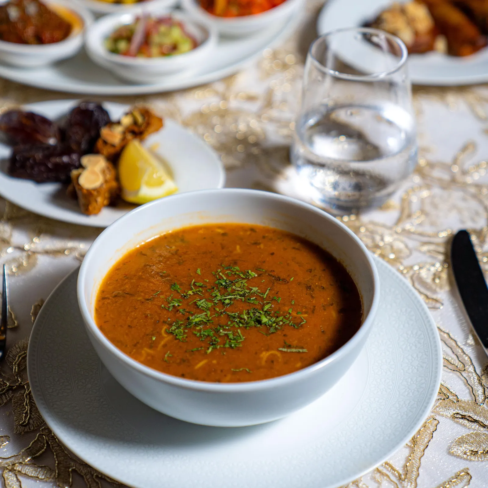 Un bol de soupe orange avec des herbes fraîches posé sur une assiette blanche, entouré de plusieurs petits plats contenant des aliments variés, le tout sur une table recouverte d'une nappe dorée