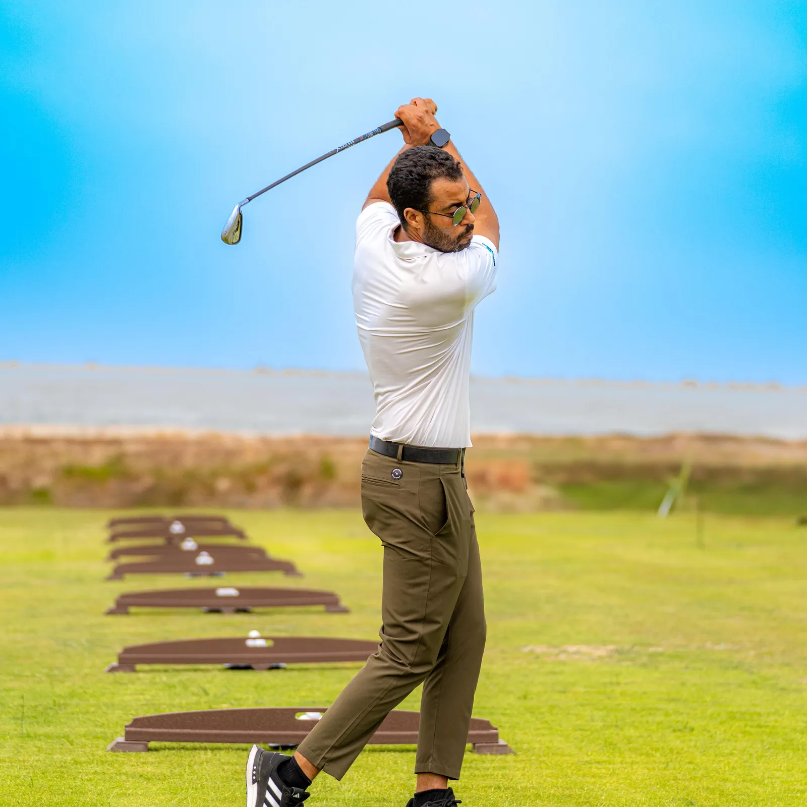 Un homme en polo blanc et pantalon vert frappe des balles de golf sur un practice en extérieur