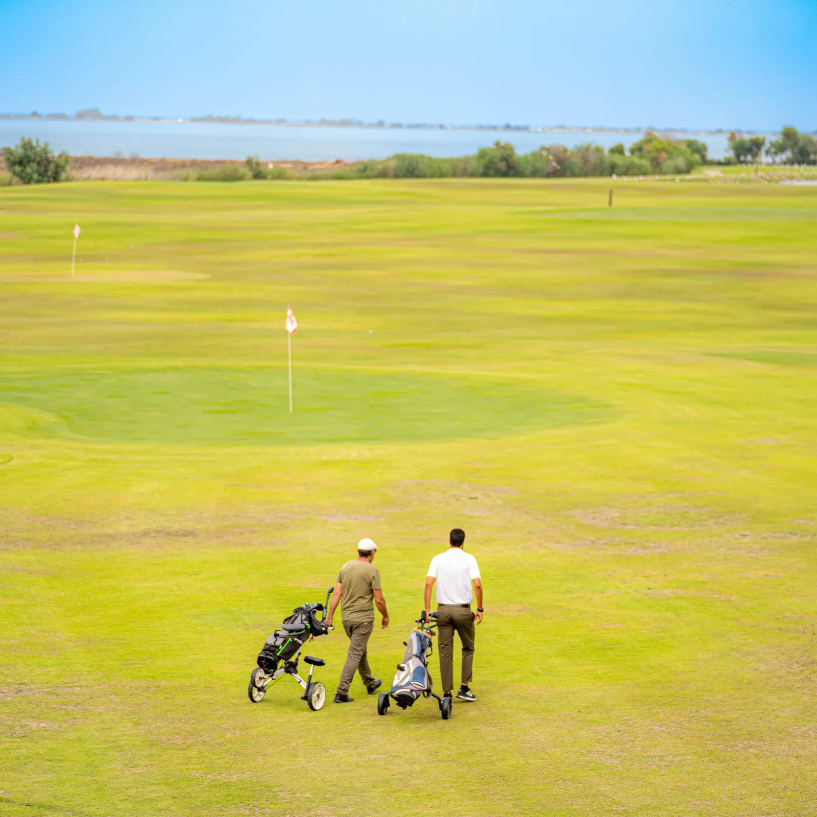Deux personnes avec des chariots de golf marchent sur un green de golf avec un drapeau visible au loin et un plan d'eau à l'arrière-plan