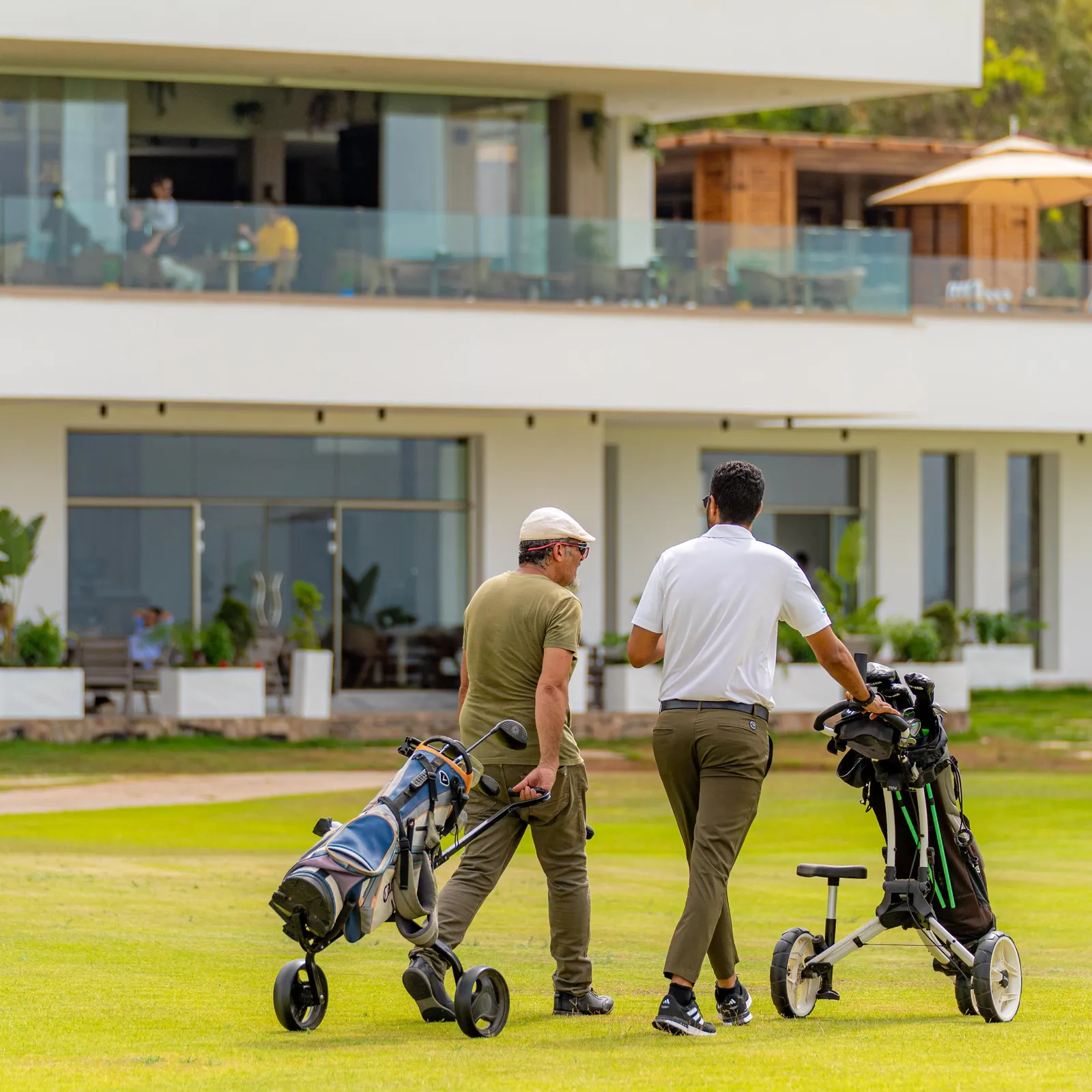 Deux hommes marchent sur un terrain de golf avec des sacs de clubs, devant un bâtiment moderne aux grandes baies vitrées