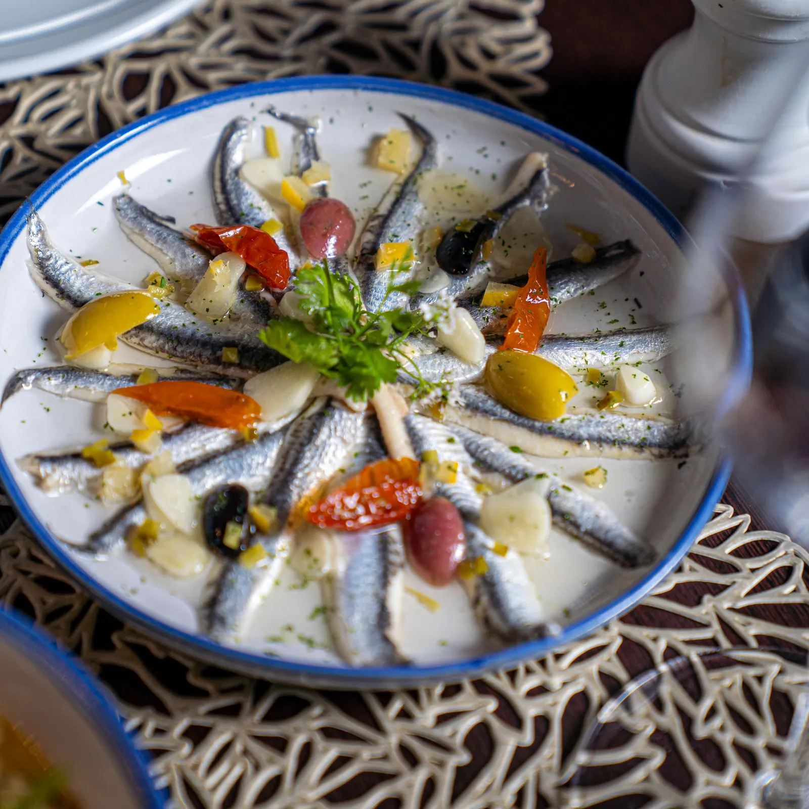Un plat de sardines grillées accompagné de légumes et d'olives, servi dans une assiette blanche à bord bleu sur une table recouverte d'une nappe à motifs