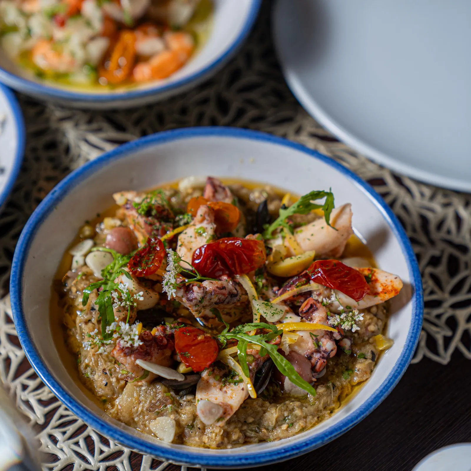 Bowl of food containing chicken, vegetables, and grains, garnished with herbs and cheese, placed on a patterned tablecloth with other bowls in the background