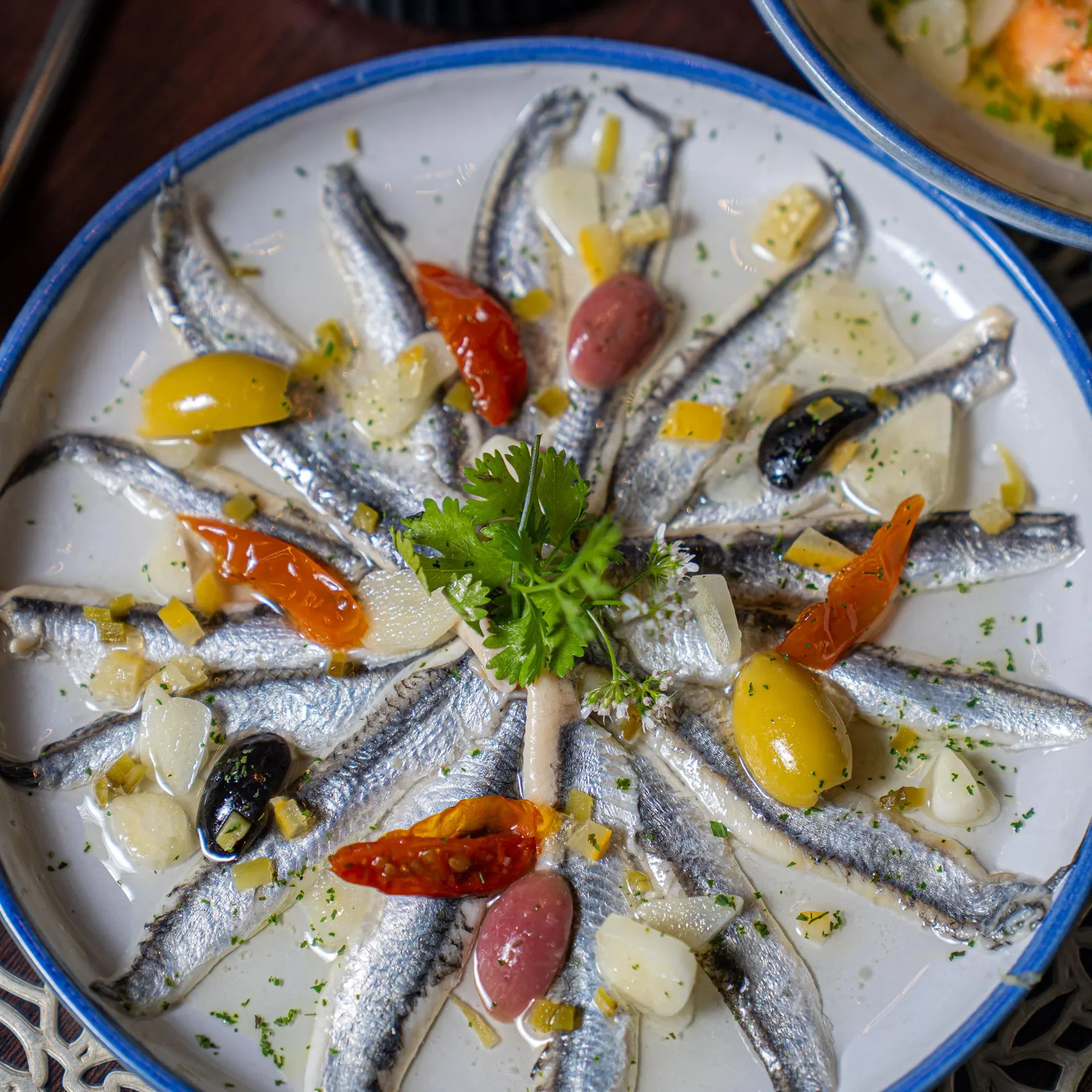 Un plat de sardines marinées avec des olives, des tomates séchées, des câpres et des herbes, présenté dans une assiette blanche à bord bleu