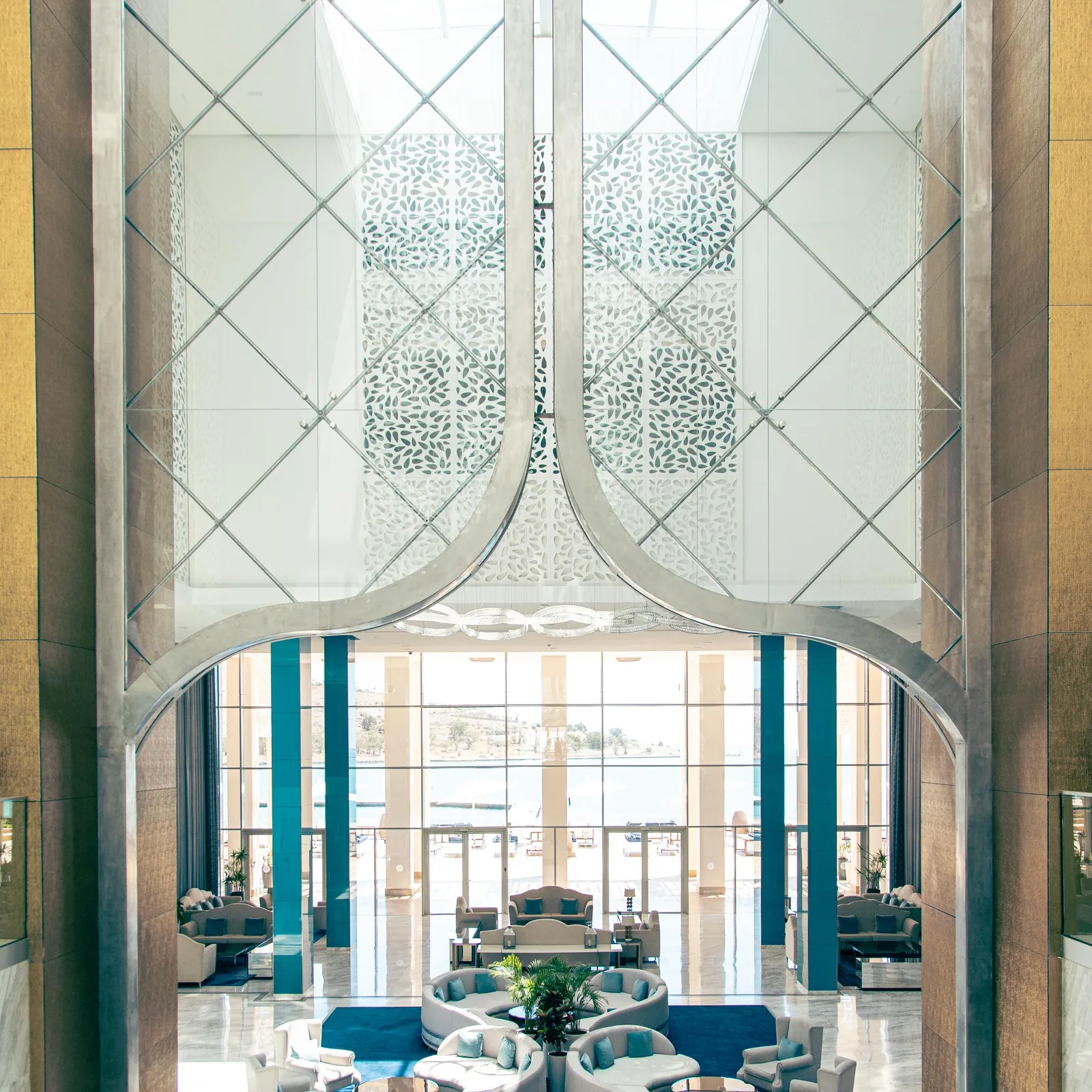 Vue d'un salon élégant à travers une structure en verre et métal avec des motifs géométriques, montrant des canapés et fauteuils disposés autour de tables basses
