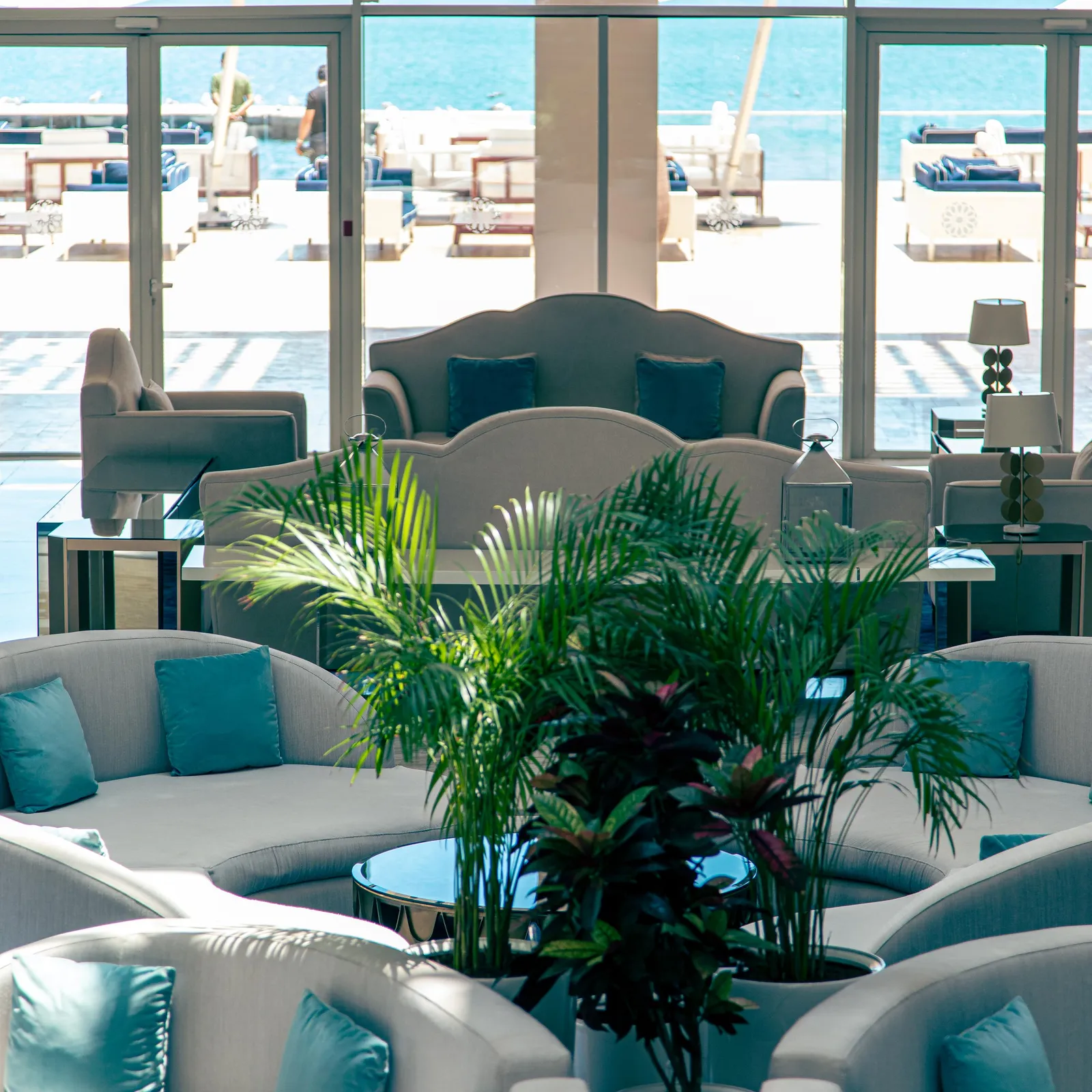 Un salon avec des canapés gris et des coussins bleus, une plante verte au centre, et une vue sur une piscine et la mer à travers de grandes baies vitrées