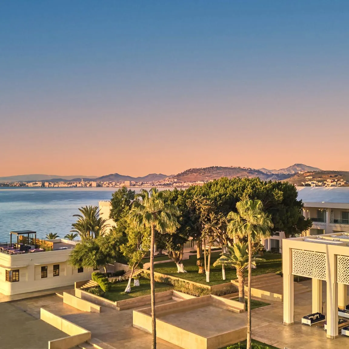 Vue d'un complexe architectural moderne en bord de mer avec des bâtiments blancs, une tour, des palmiers et une mer calme au coucher du soleil