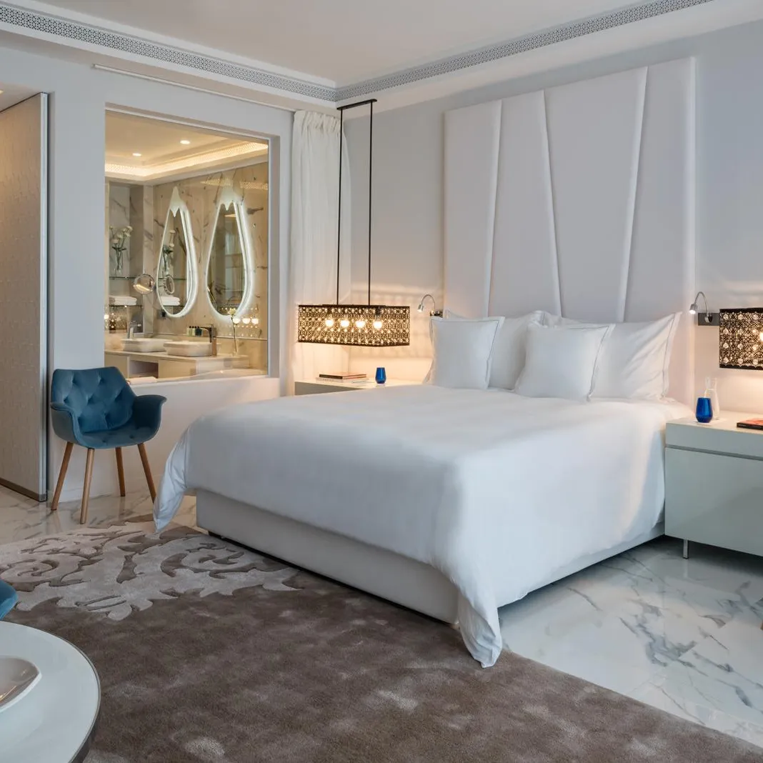 Chambre luxueuse avec lit blanc, deux fauteuils, table de chevet et vue sur une salle de bain avec miroirs ovales