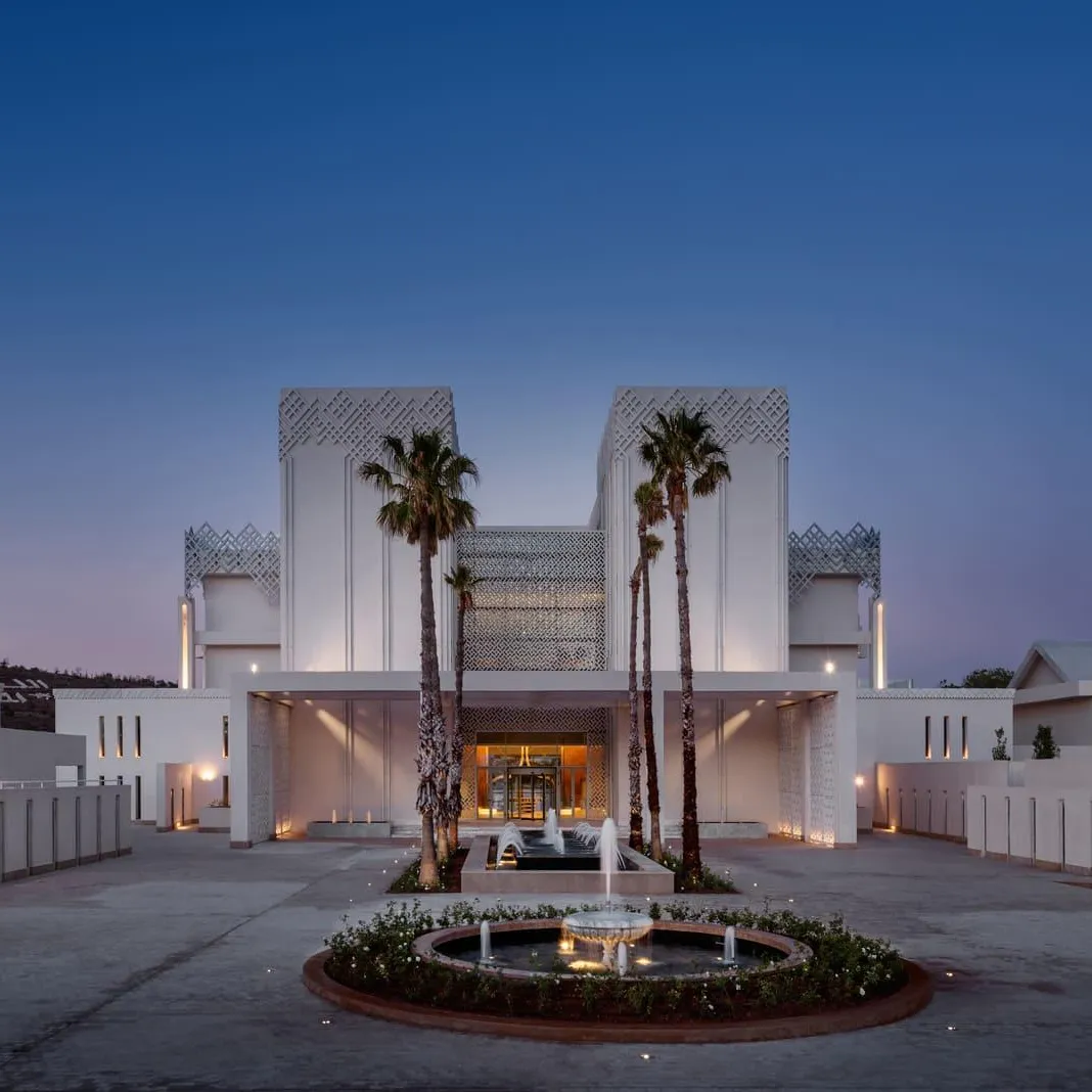Bâtiment moderne avec façade blanche et motifs géométriques, entouré de palmiers et de fontaines, éclairé à la tombée de la nuit