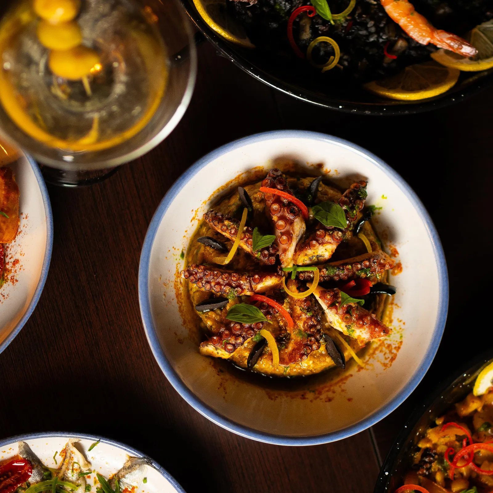 Un plat de calamars grillés avec des légumes et des herbes, accompagné d'autres assiettes de fruits de mer et d'une boisson dans un verre
