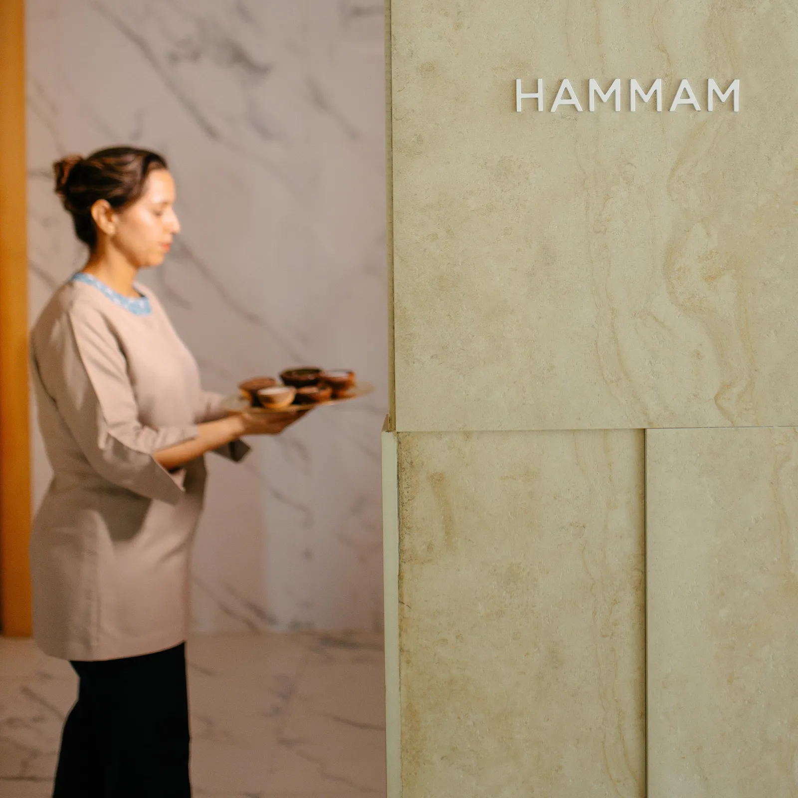 Femme en tenue traditionnelle portant un plateau dans un intérieur aux murs de pierre avec l'inscription "HAMMAM"