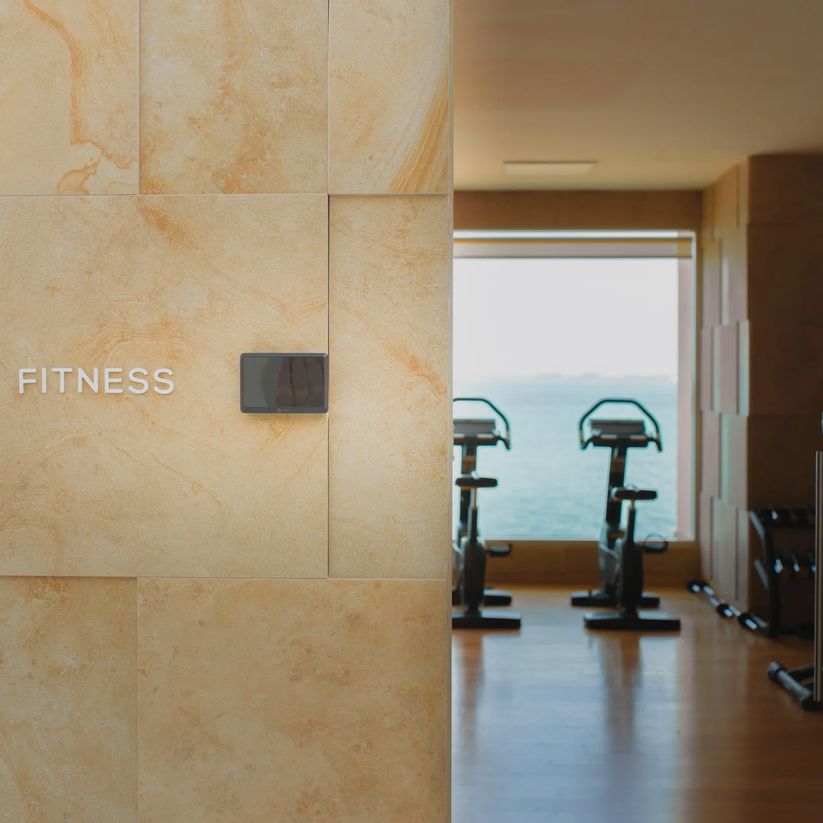 Vue d'un espace de fitness avec des vélos d'exercice, visible à travers une porte ouverte, et un mur en pierre avec l'inscription "FITNESS"