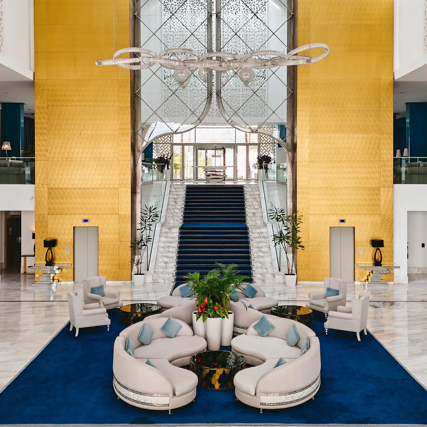 Lobby d'hôtel avec des canapés blancs, des plantes vertes, un escalier central et un grand lustre suspendu