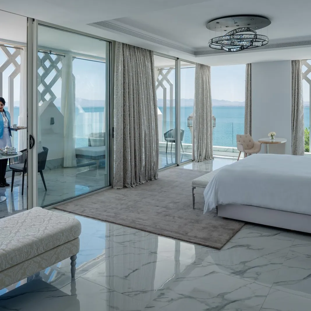 Chambre d'hôtel luxueuse avec vue sur la mer, un lit, un canapé, une table à manger et une personne servant un plateau