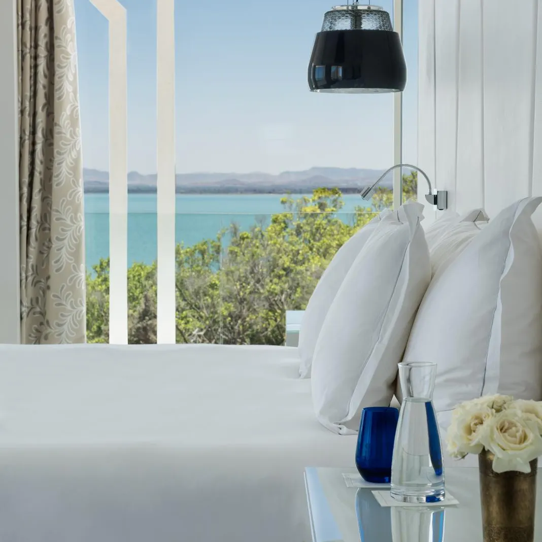 Vue d'une chambre avec un lit blanc et des coussins, une table de chevet avec des vases et des fleurs, et une grande fenêtre donnant sur une mer bleue avec des montagnes au loin