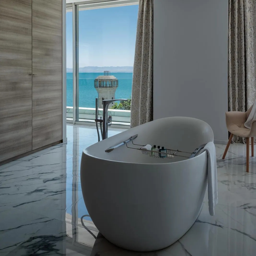 Baignoire blanche avec des serviettes posées dessus, située dans une pièce avec vue sur la mer et un fauteuil recouvert d'une serviette