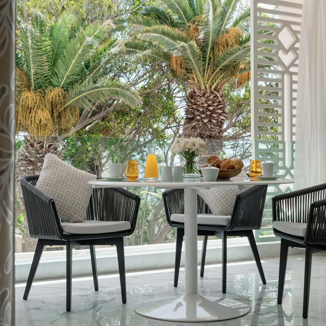 Table ronde avec quatre chaises en rotin noir et coussins gris, vue depuis une porte ouverte, avec des plantes et des éléments décoratifs sur la table en arrière-plan
