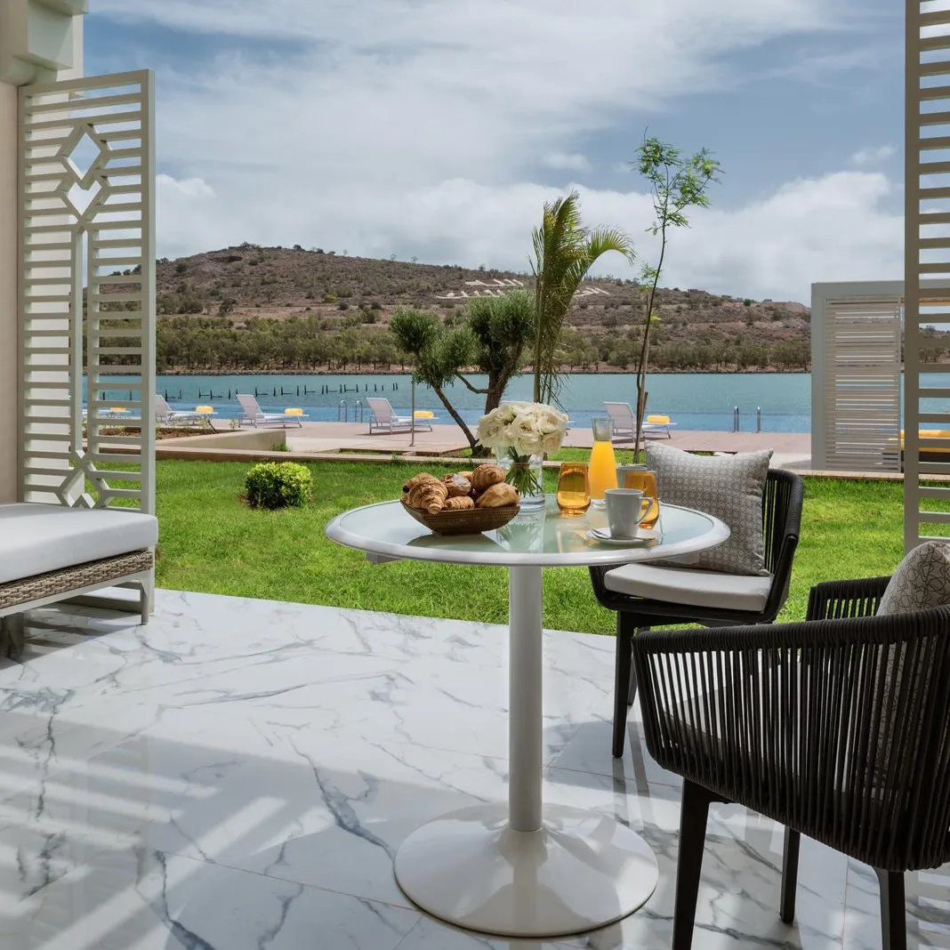 Terrasse avec table, chaises et canapé donnant sur une pelouse et un lac avec des transats et des montagnes en arrière-plan