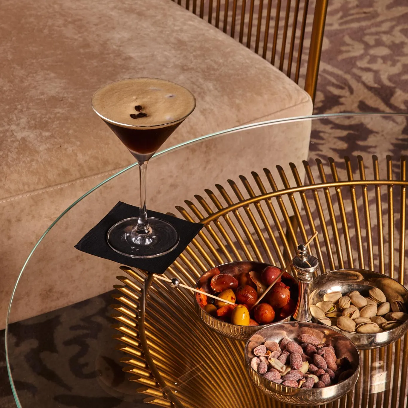Un verre de cocktail posé sur une table basse en verre et métal doré avec des bols de fruits secs et de pistaches